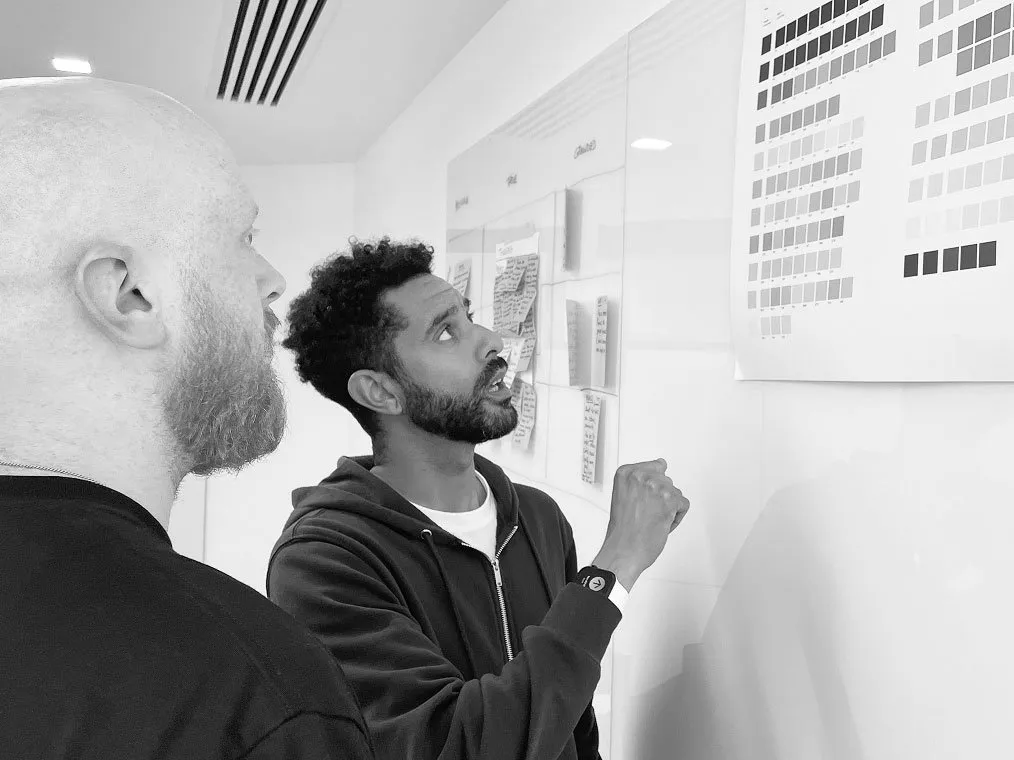 Two men examine a large chart of design swatches posted on an office wall.