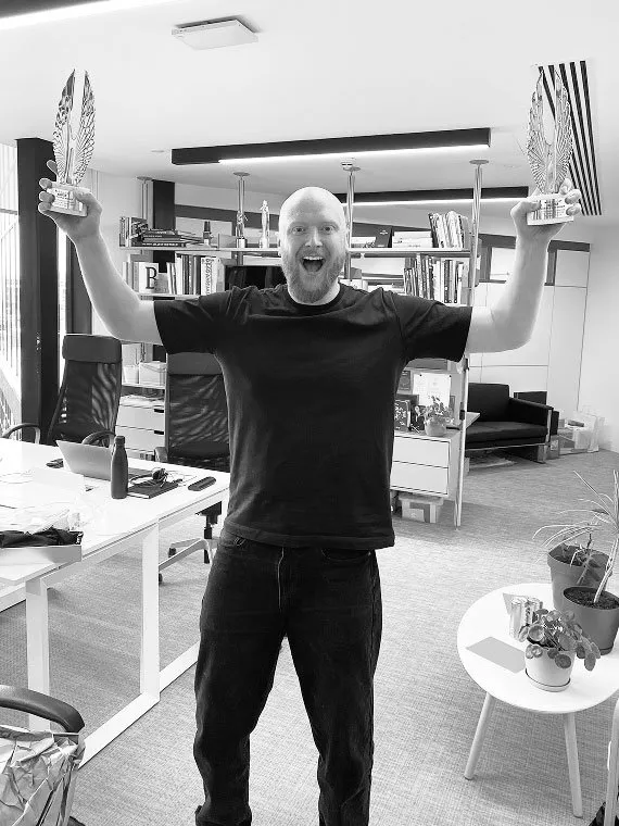A smiling bald man with a beard stands in an office, excitedly holding up two silver wing-shaped trophies with raised arms.