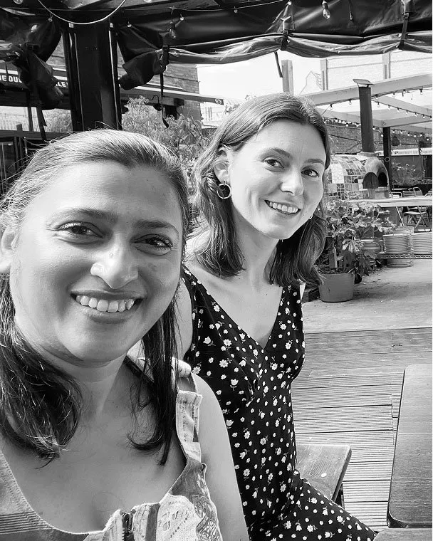 Black and white selfie of two smiling women sitting at an outdoor restaurant patio with a wooden deck and plants in the background.