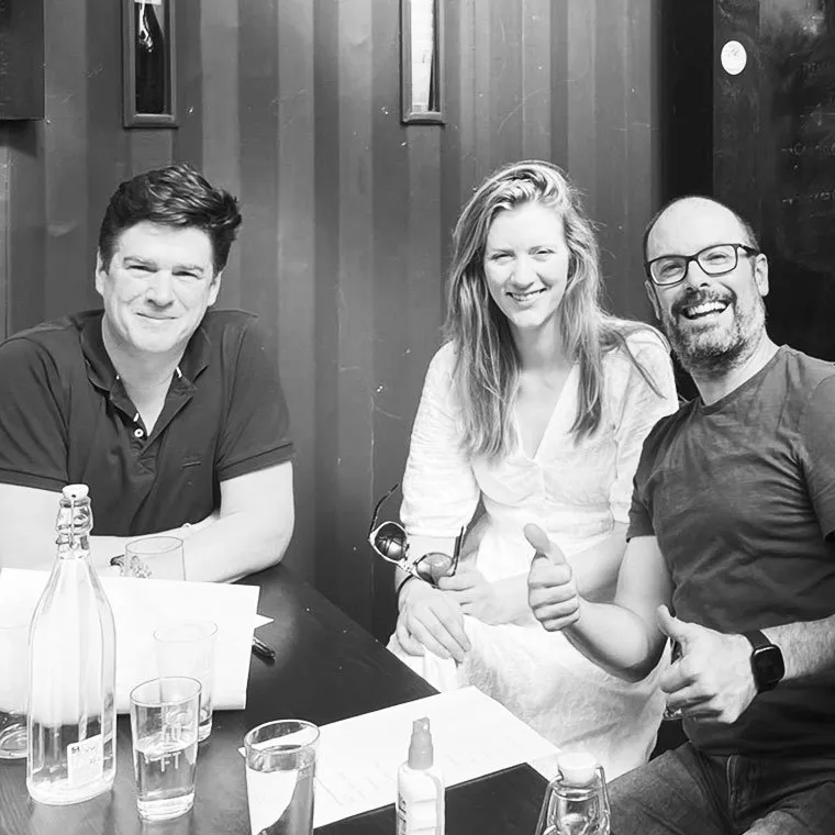 A black and white photo of three smiling people sitting together at an outdoor table, with the man on the right giving two thumbs-up.