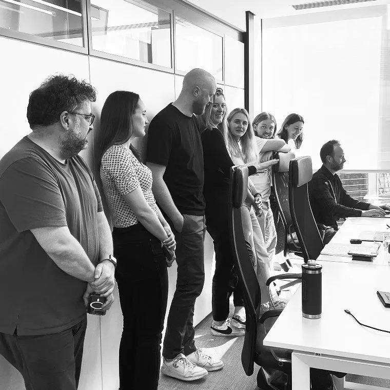 Black and white photo of a group of coworkers standing in a row against an office wall, looking toward a colleague seated at the end of a long conference table.