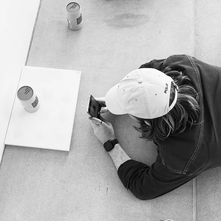 Overhead black and white view of a person lying on a concrete surface, using a smartphone to photograph cylindrical product containers placed on a white board.
