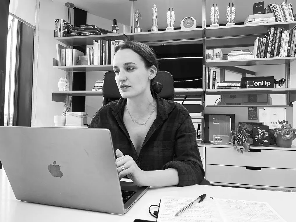 A woman sits at an office desk working on a laptop, with a notebook and pen nearby and modern shelves filled with books and awards behind her in black and white.