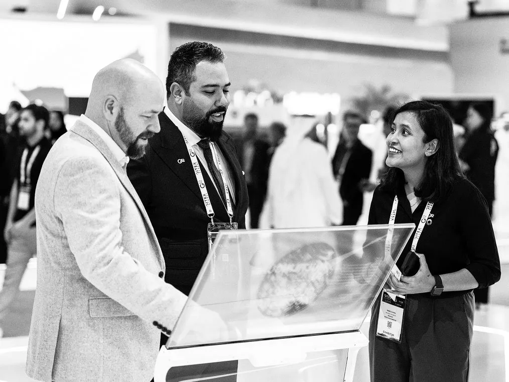 A black and white photo of three professionals gathered around a transparent interactive display screen at an exhibition.