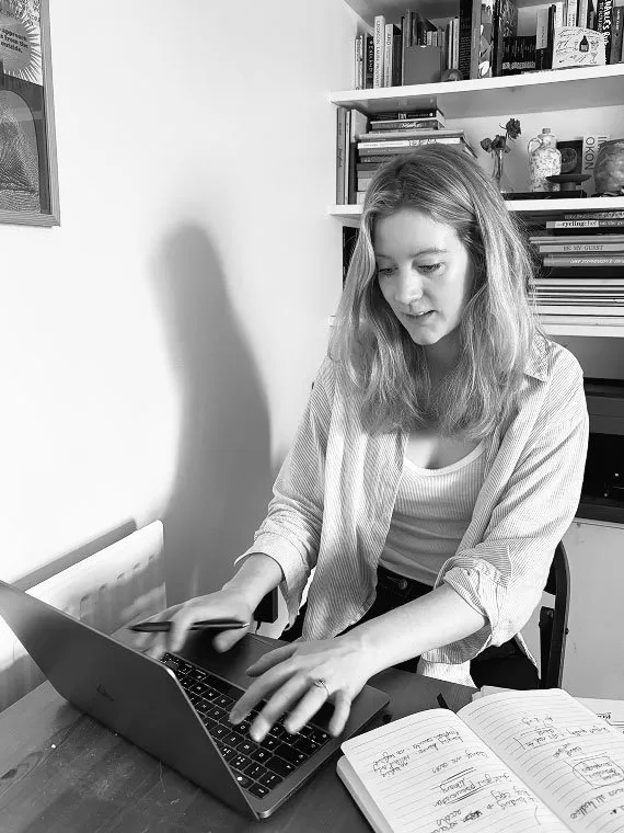 Black and white photo of a woman sitting at a desk typing on a laptop with an open notebook beside her, in front of bookshelves filled with books.