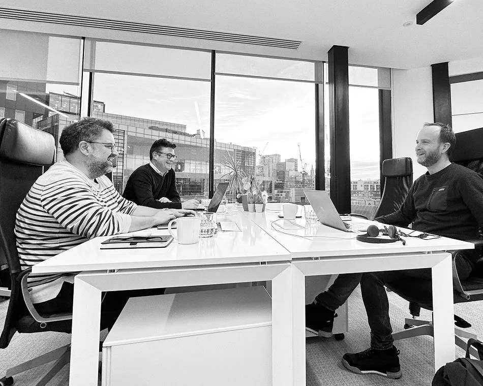 Three men smile and converse while seated around a white desk in a modern office with large windows overlooking a city skyline.