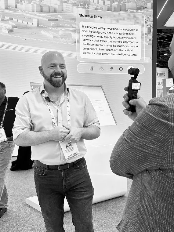 A smiling man wearing an exhibitor badge stands at a technology trade show booth while being filmed with a handheld gimbal camera. In the background is a digital display presenting information about infrastructure and connectivity.