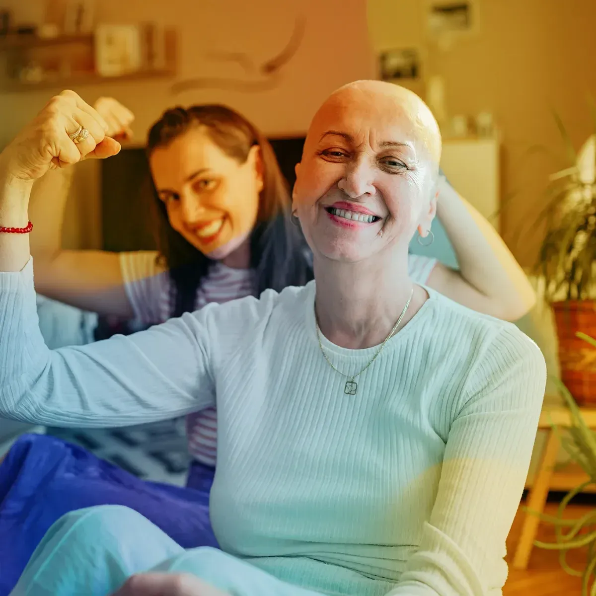 An older woman with a shaved head smiles and flexes her arm while a younger woman laughs with her arm around her, sitting together at home.