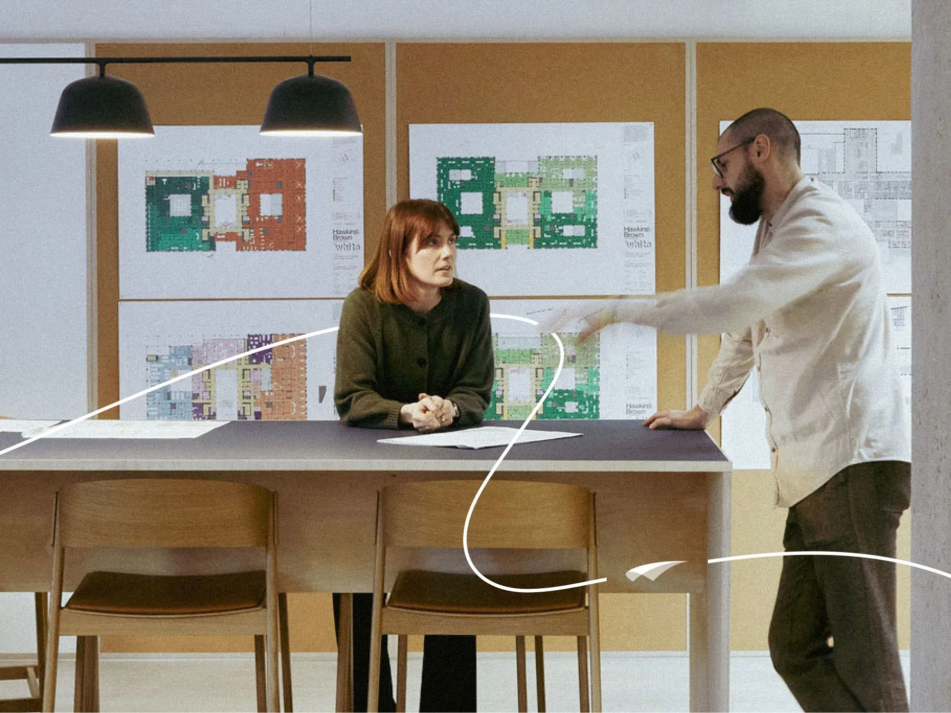 Two colleagues in an architecture office discussing a project at a high table, with colorful floor plans pinned to the wall behind them and an illustrated paper airplane trail looping across the scene.